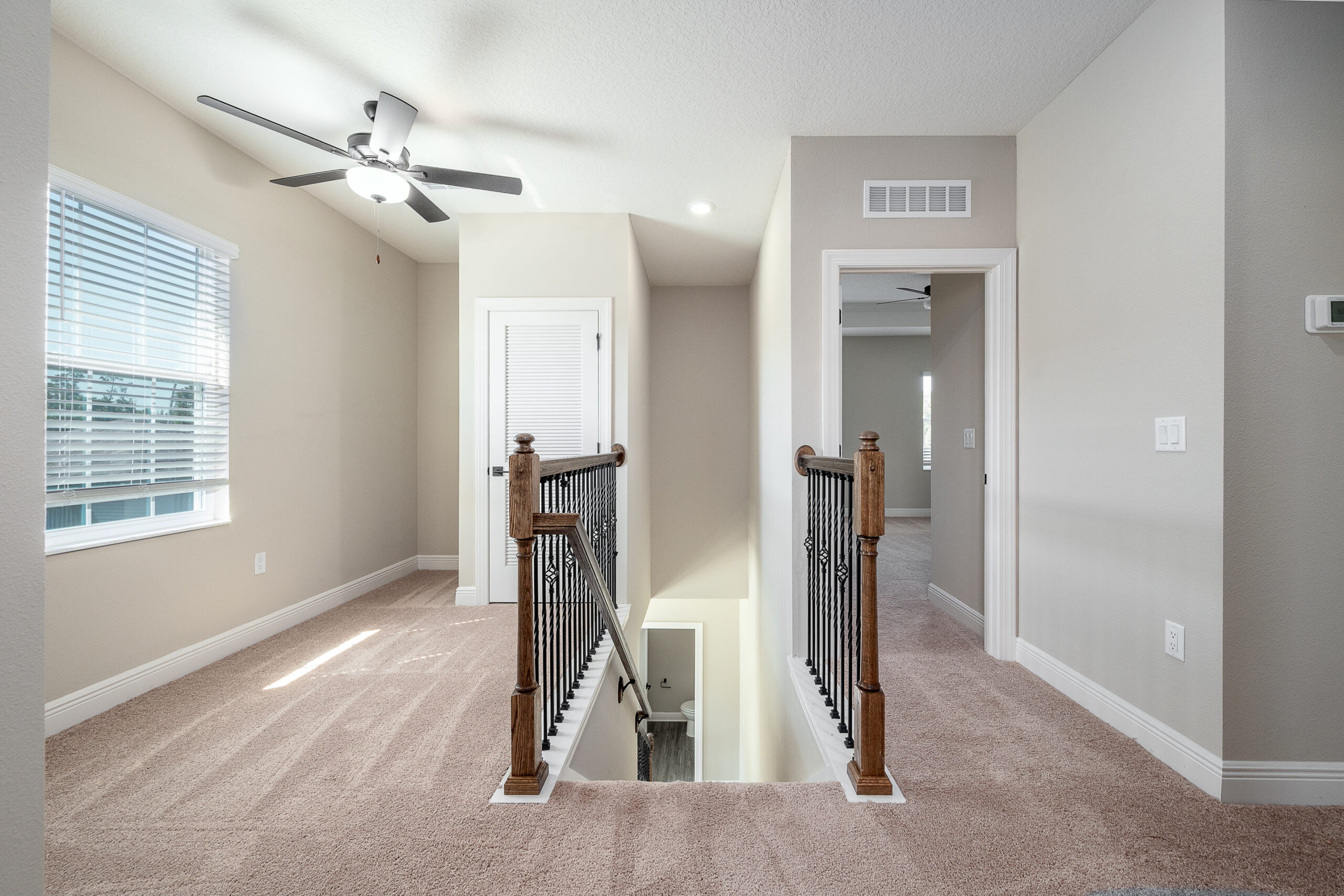This photo shows a carpeted upstairs landing with a wooden and wrought iron railing. A ceiling fan hangs above, and there's a window to one side. The area has two white doors, one leading downstairs, and the other presumably to a room. Natural light brightens the space.