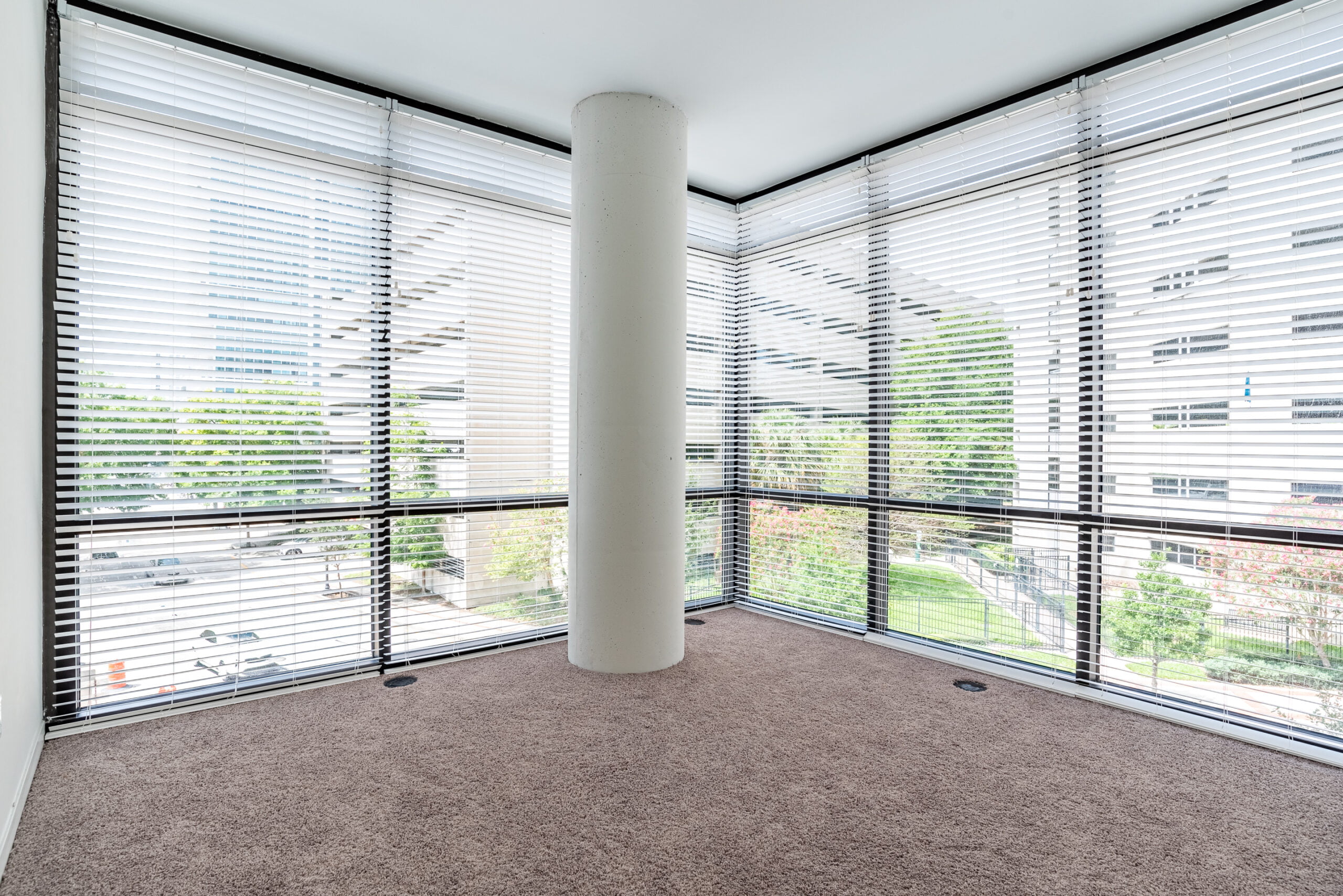 This photo features a room with a large column in the center, wall-to-wall windows covered by horizontal blinds, and a carpeted floor, providing an unobstructed urban view.