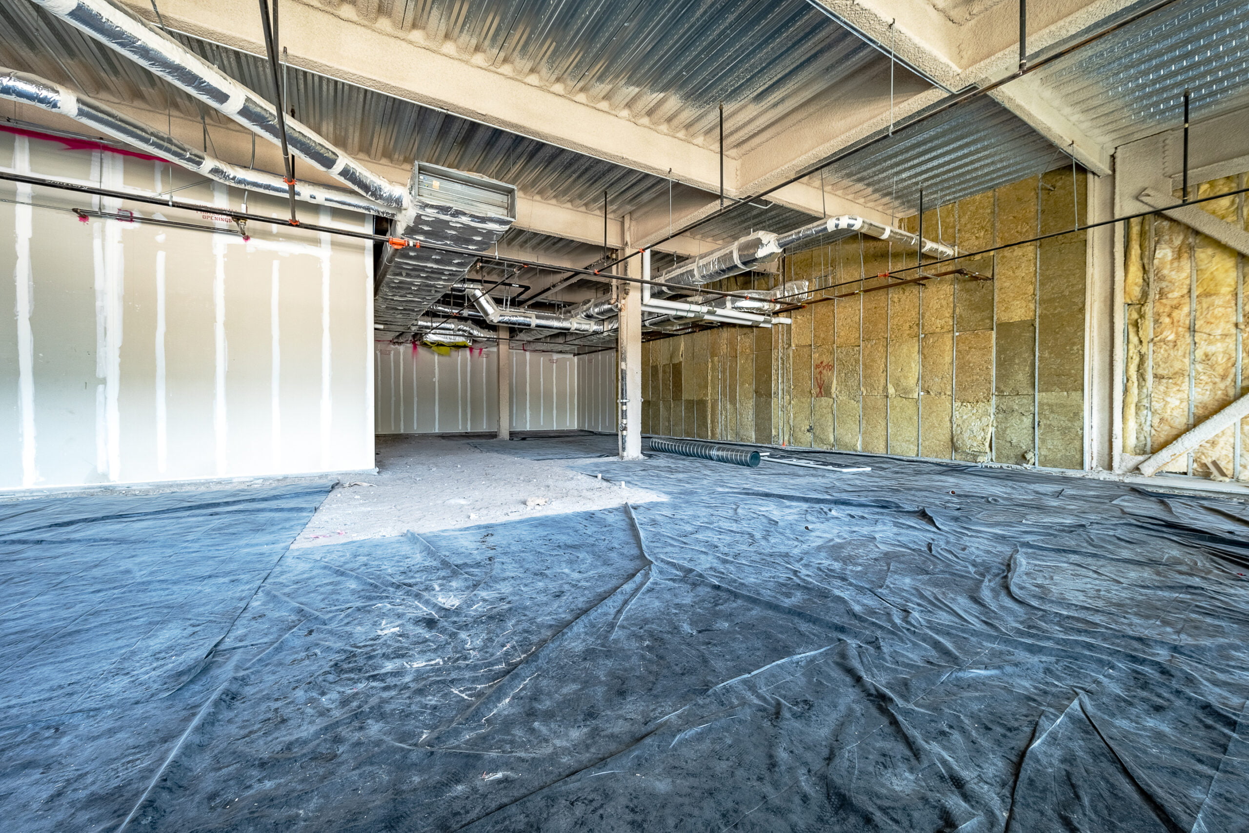 This photo shows an unfinished interior space under construction, with exposed insulation, ductwork, and structural elements. A protective plastic sheet covers the floor.