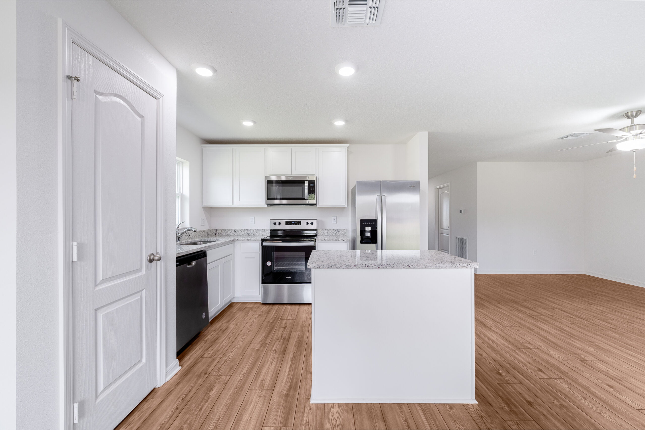 This photo shows a bright, modern kitchen with white cabinetry, stainless steel appliances, and a central island. The space opens into a larger room with wooden flooring and a white ceiling with recessed lighting, conveying a clean, open-plan living area.