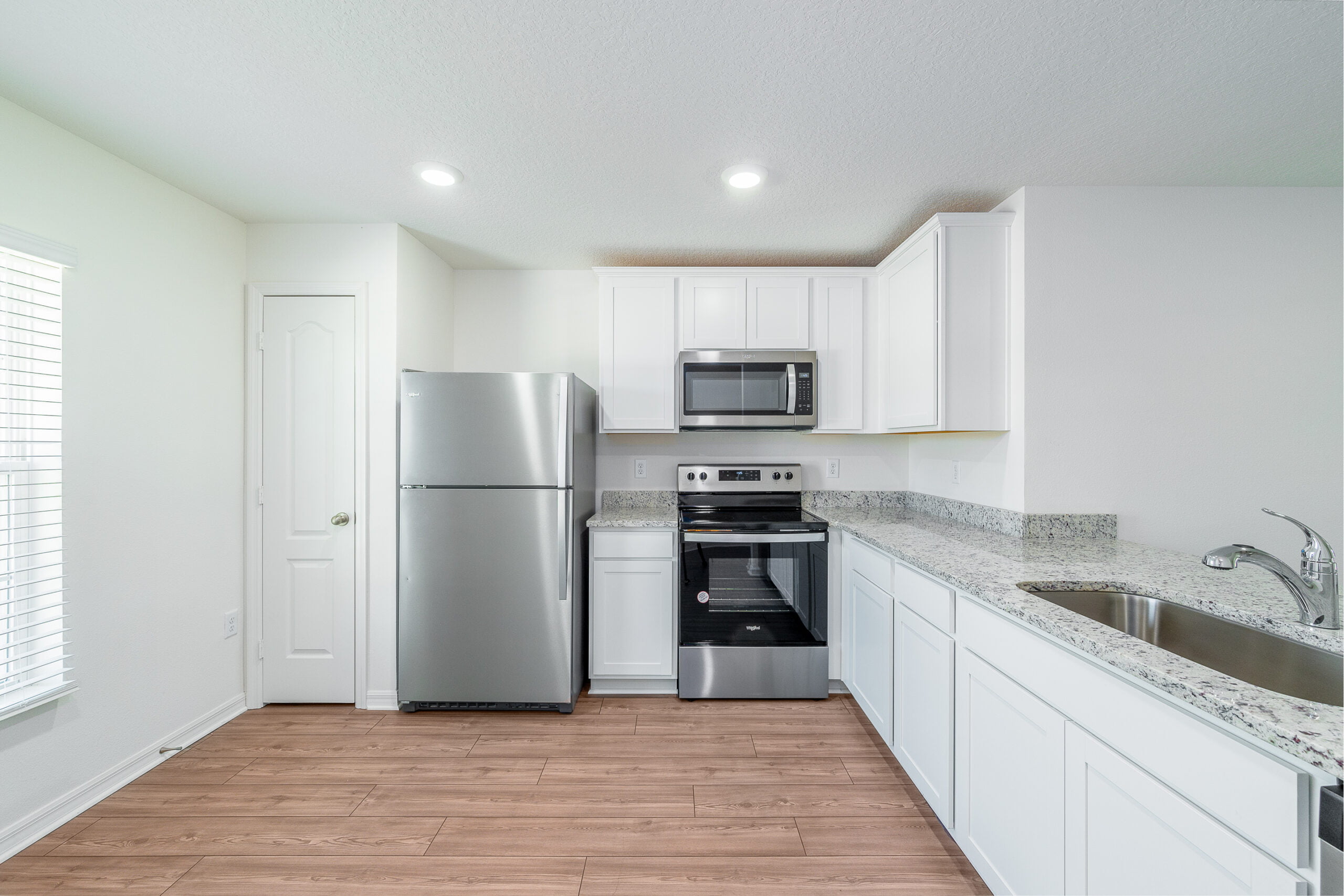 This photo showcases a modern kitchen with white cabinetry, stainless steel appliances, granite countertops, and recessed lighting. The flooring is a laminate wood, and natural light filters in through the blinds. The space is clean and uncluttered, suggesting a new or recently updated interior.