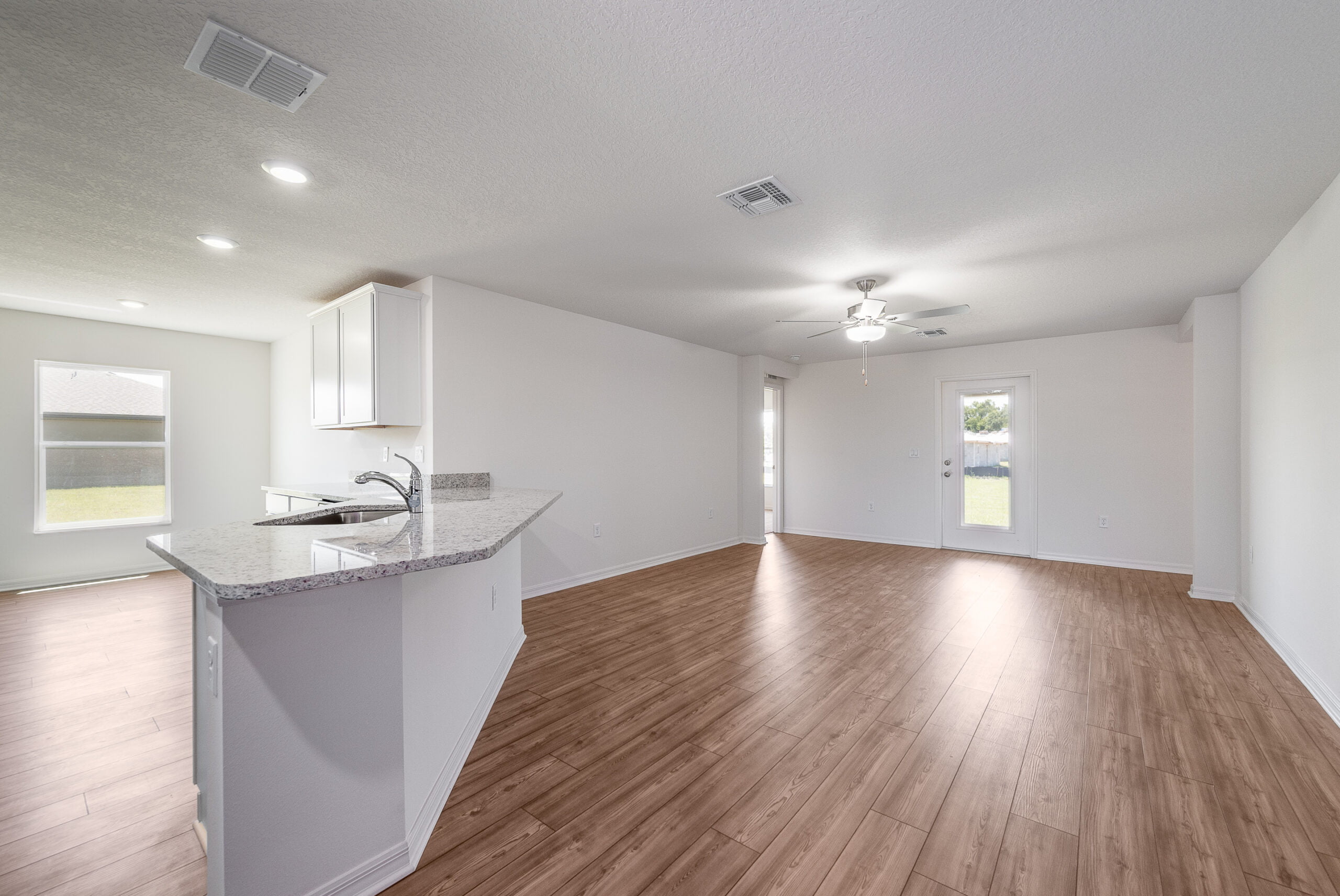 This photo displays an empty, bright room featuring a kitchen area with modern appliances and a breakfast bar. The space has laminate wood flooring, recessed lighting, and a ceiling fan, with natural light entering through windows and a glass door leading to a backyard.
