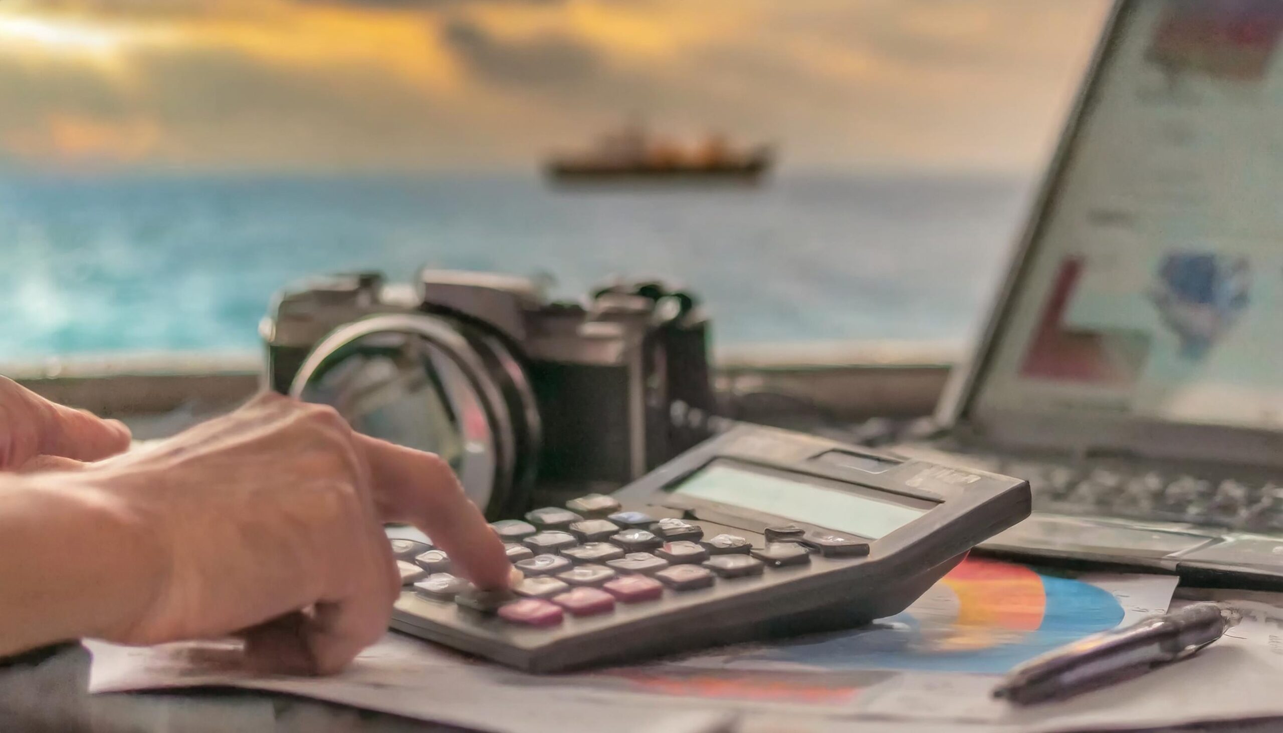 Workspace during sunset with a calculator, camera, and laptop on a desk, symbolizing commercial photography planning with an ocean view.
