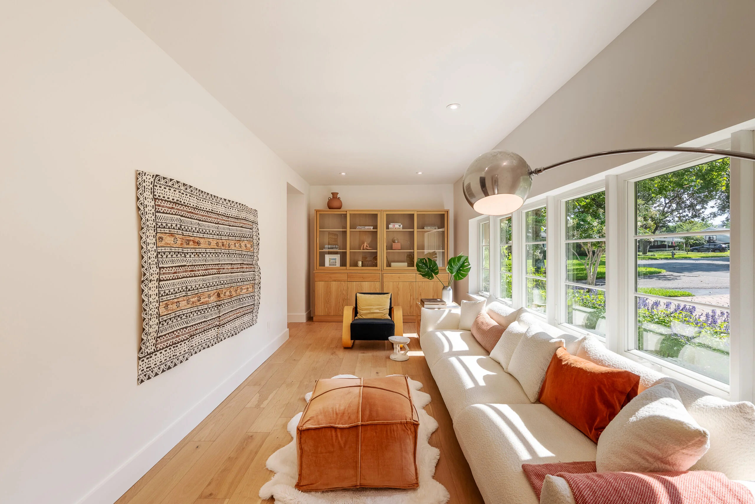 Modern Bright Living Room with Elegant Decor and Natural Light Modern living room with light wood flooring, a large cream sofa with throw pillows, an orange pouf on a fluffy rug, an arc-shaped floor lamp, a decorative textile wall hanging, and a wooden cabinet with glass doors.